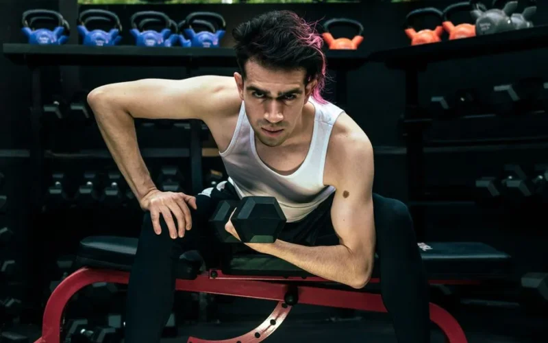 A man intensely lifting a dumbbell on a bench in a gym environment.