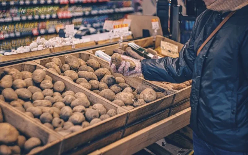 A shopper picks potatoes from a wooden crate in a grocery store produce section.