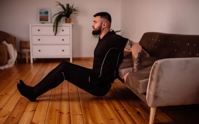 A bearded man in black fitness attire exercising at home, showcasing arm tattoos and a healthy lifestyle.