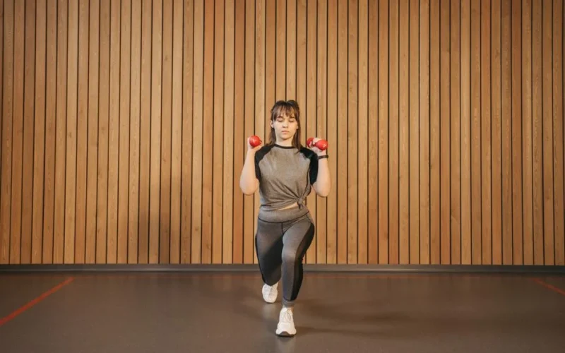 A woman performing lunges with dumbbells in a modern indoor gym setting.