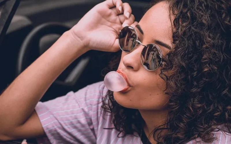 Woman with curly hair and sunglasses blowing gum in car, exuding style and confidence.