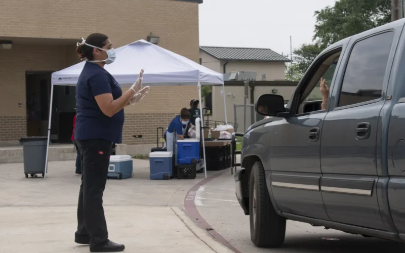 Harlandale Independent School District's (HISD) Armando Leal Jr. Middle School Child Nutrition staff Laura Mares greets a driver and confirms the number of children who need meals from U.S. Department of Agriculture (USDA) Food and Nutrition Service (FNS) National School Lunch Program (NSLP) Seamless Summer Option's (SSO) alternative to traditional congregant feeding; this affords safe distancing with curbside distribution during the COVID-19 pandemic in the neighborhoods supported by HISD, in San Antonio, TX, on April 9, 2020. SSO has less paperwork, making it easier for schools to feed children during the traditional summer vacation periods and, for year-round schools, long school vacation periods, and during unanticipated school closures (i.e.the current coronavirus emergency). For more information about Seamless Summer and Other Options for Schools, please go to fns.usda.gov/sfsp/seamless-summer-and-other-options-schools. Here, a greeter welcomes drivers and ask how many children are being fed. The amount is called out to teammates who then place the bagged meals of hot lunches and cold breakfasts, on a table, then steps back. Drivers lower their windows and pick-up the meals from the table. This alternate workflow with a 6-person team of child nutrition staffers delivers more than 500 meals in 90 minutes. The school has received many thank-you messages from parents and students for providing this service. The staff are highly motivated, happy to see, wave to, and serve the children they have seen grow up. Because this site is closed tomorrow, children are given two days' worth of breakfast and lunch meals. The meals fulfill USDA standards and include USDA commodities of turkey sandwiches and apple sauce/strawberry cups. Because of the portable needs of the meals, a bag of multigrain chips replaced hummus for today's grains. The hot meal includes a corndog made from a USDA recipe. Because of the portable needs of the meals, a bag of multigrain chips replaced hummus for today's grains. For more information about HISD Child Nutrition Program, please go to harlandale.net/cms/one.aspx?portalId=829982&pageId=1745444. The National School Lunch Program (NSLP) is a federally assisted meal program operating in public and nonprofit private schools and residential child care institutions. It provides nutritionally balanced, low-cost, or free lunches to children each school day—the National School Lunch Act, signed by President Harry Truman in 1946. For more information about NSLP, please go to fns.usda.gov/nslp/nslp-fact-sheet. Children need good nutrition all year long. The Summer Food Service Program, National School Lunch and Breakfast Programs, and the Seamless Summer Option make it possible for schools to provide nutritious meals after the regular school year ends. For more information about FNS, please go to fns.usda.gov USDA Photo by Lance Cheung.