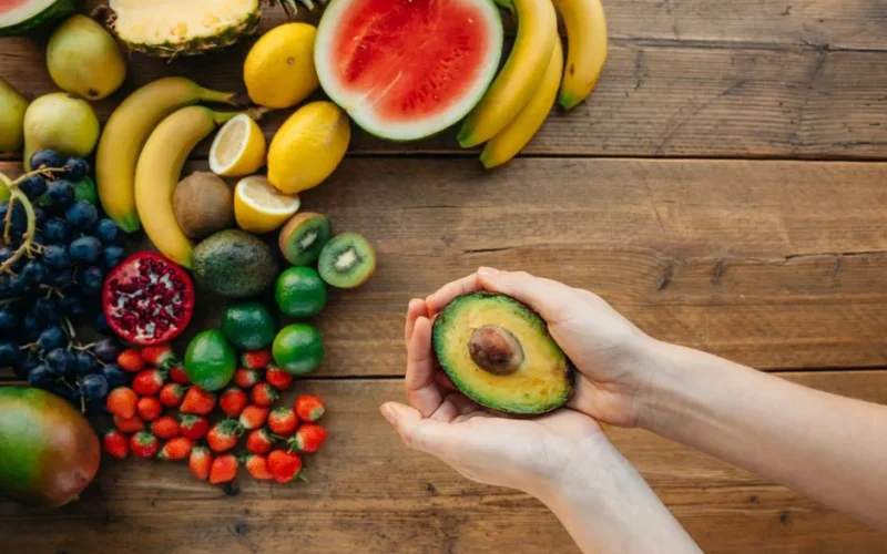 Hands holding a halved avocado with a colorful assortment of fruits on a wooden table.