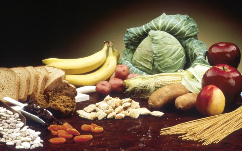 A wooden table containing: a ladle full of beans, a sliced loaf of brown bread, a bunch of bananas, muffins, small potatoes, a head of cabbage, an ear of corn, a pile of cereal, yams, apples, a nectarine and some spaghetti. See also AV-3905 and AV-3906. AV Number: AV-8812-3429 Reuse Restrictions: None - This image is in the public domain and can be freely reused. Please credit the source and/or author listed above.