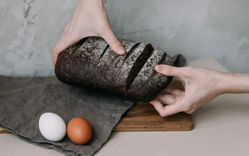 Hands slicing dark rye bread with eggs on a wooden board, perfect for breakfast themes.