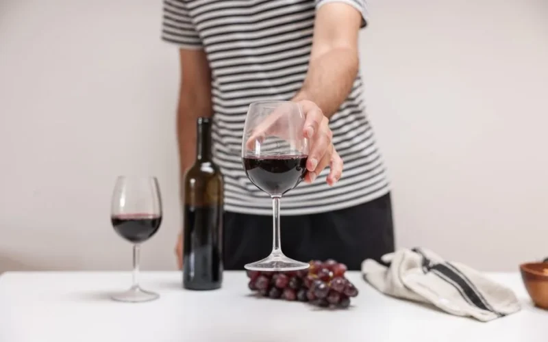 A person in a striped shirt holding a glass of red wine next to grapes and a wine bottle.