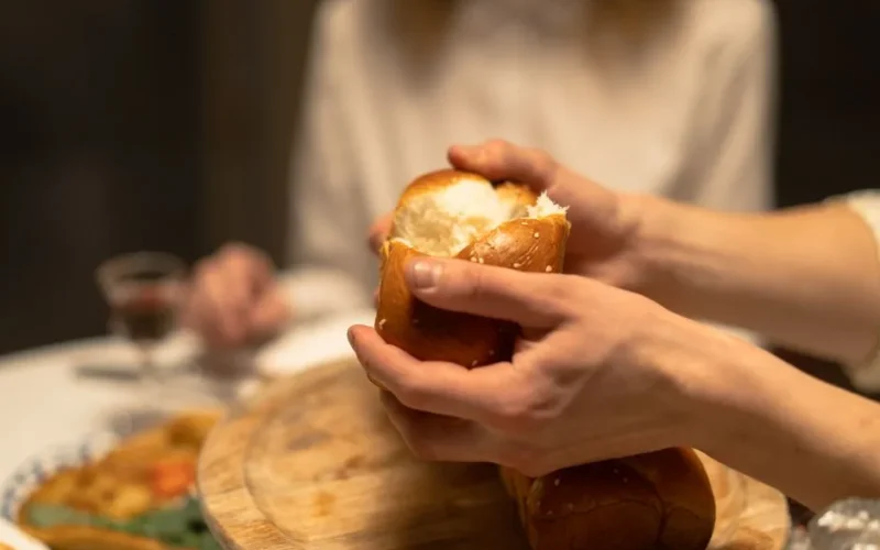 Close-up of hands breaking challah bread at a festive indoor dinner setting.