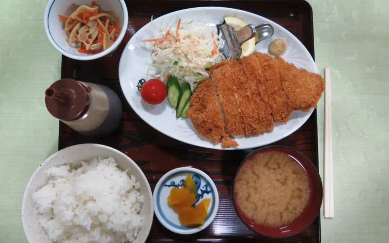 A tonkatsu (fried breaded pork cutlet) dinner served in Akita, Japan. On the tray are the cutlet, cabbage, cucumbers, tomatoes, lotus root, carrots, rice, pickled burdock, sauce, and a bowl of miso soup.