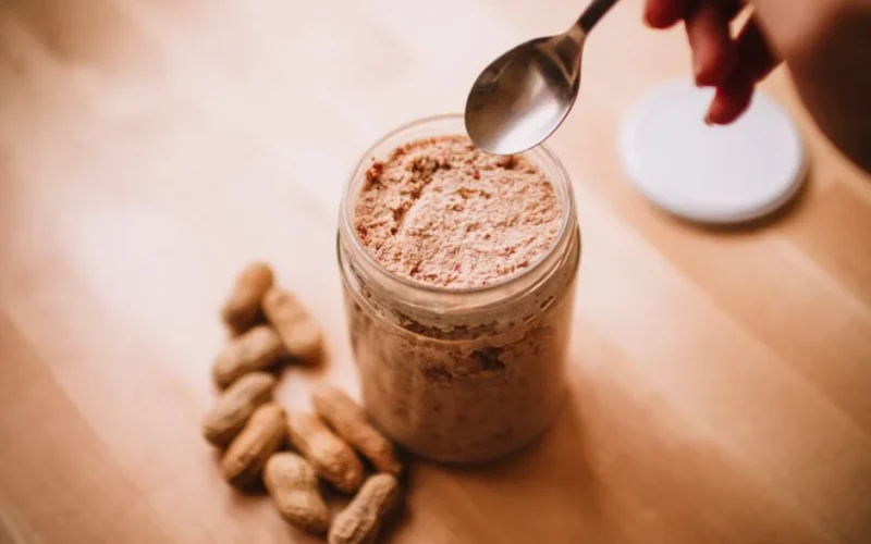 A glass jar of homemade peanut butter on a wooden table with peanuts and a spoon.