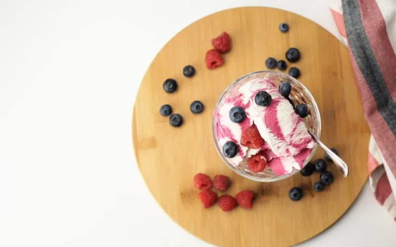 A high-angle shot of berry vanilla ice cream garnished with blueberries and raspberries on a wooden board.