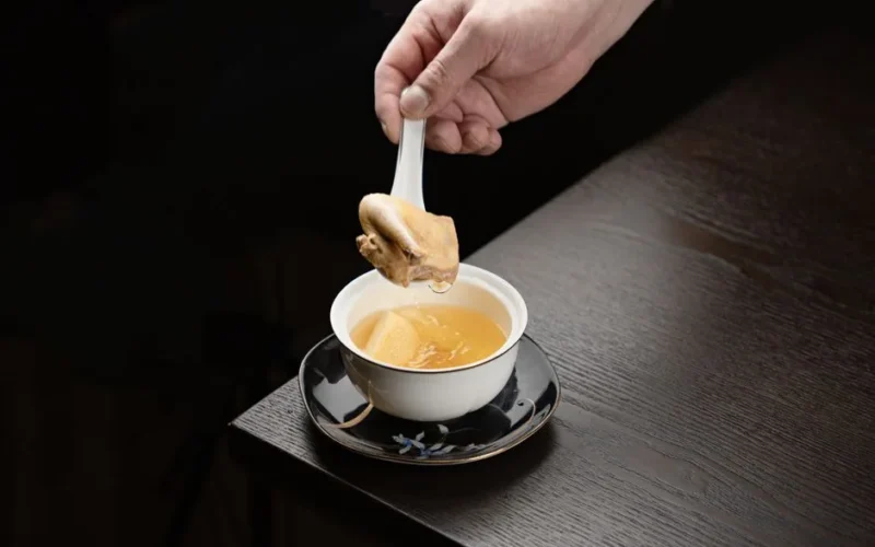 Close-up of a hand holding a spoon with chicken in a bowl of rich broth on a wooden table.