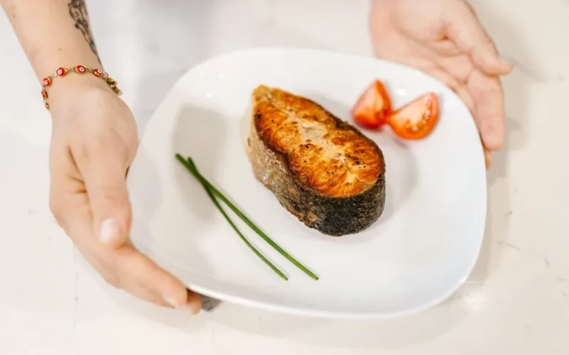 Close-up of grilled salmon served with tomatoes and chives on a plate, held by tattooed hands.