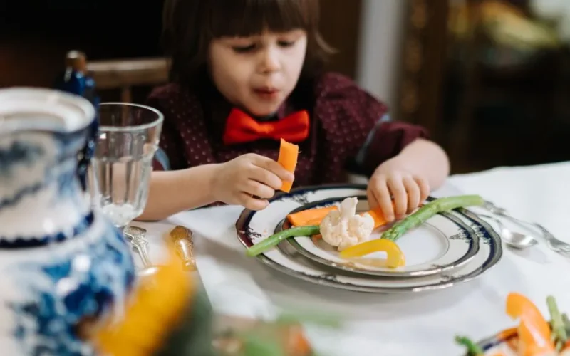 A child in a red bow tie enjoys a healthy meal with vegetables indoors.