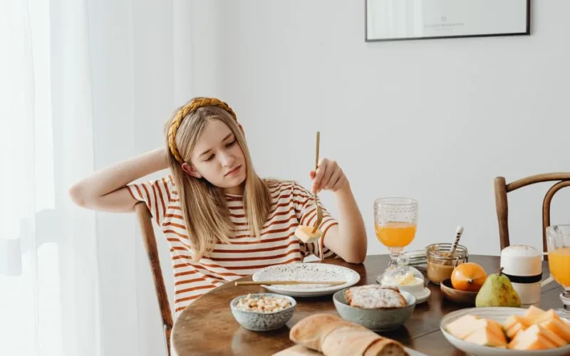 Teen girl in striped shirt at breakfast table with fruits, bread, and juice. Casual morning setting.