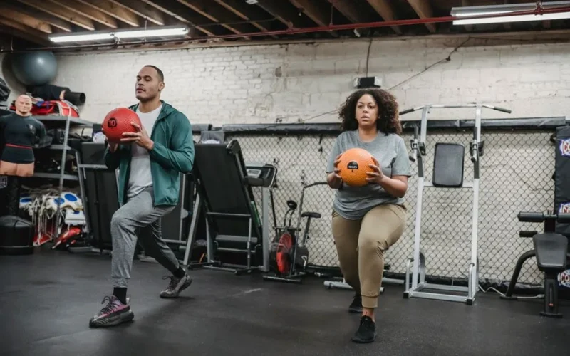 Full body of focused overweight African American female with fit ethnic male trainer doing forward lunges with medicine balls during functional workout in gym