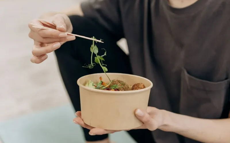 A woman holding a paper bowl filled with a healthy vegetarian meal in an indoor setting, promoting wellness.