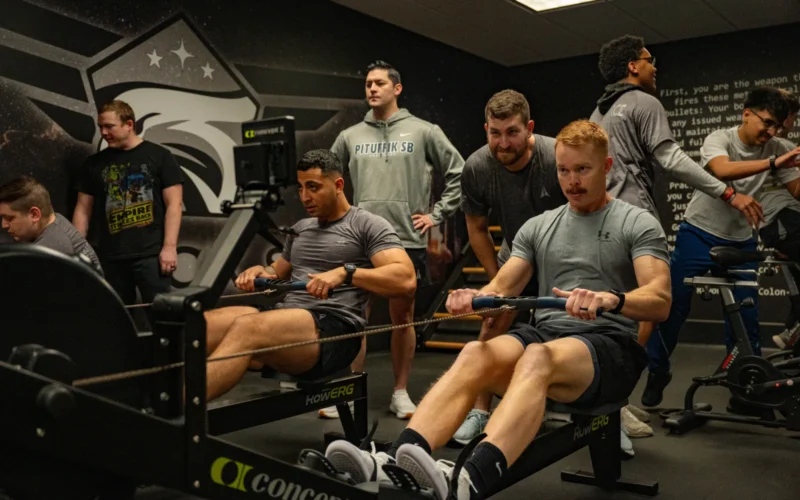Positive young obese tattooed female in activewear sitting on rowing machine and doing pulling exercise during intense training with Asian male personal coach in modern gym