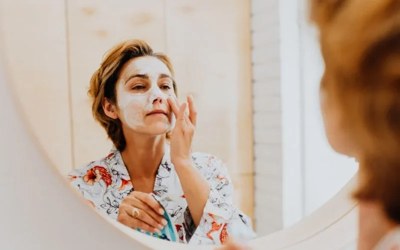 A woman applying a cosmetic mask for skincare in front of a mirror.