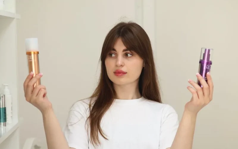 A young woman compares two cosmetic products while standing indoors, unsure of which to choose.
