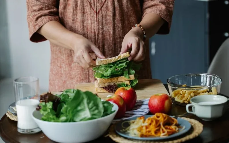 Unrecognizable female cook putting slice of bread on green lettuce while standing at table with assorted food and making breakfast