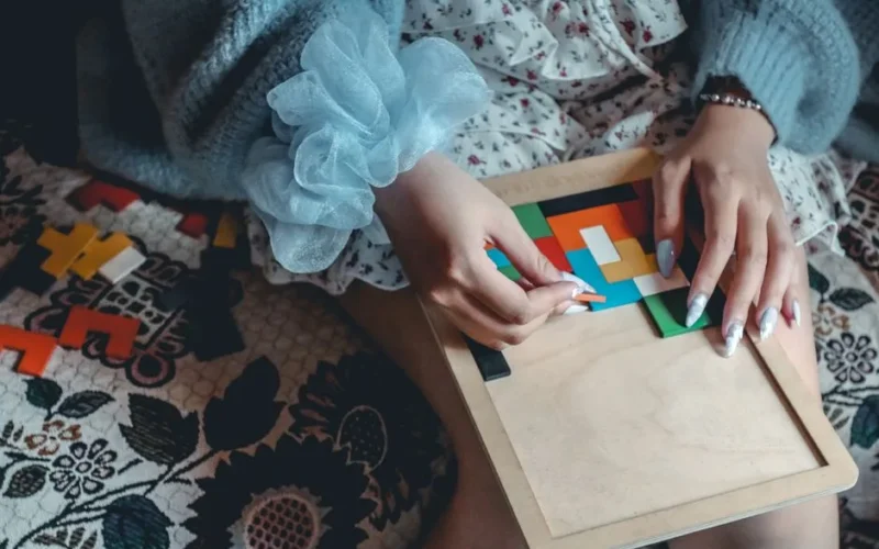 A woman engaged in solving a colorful puzzle indoors, wearing a blue sweater and floral dress.
