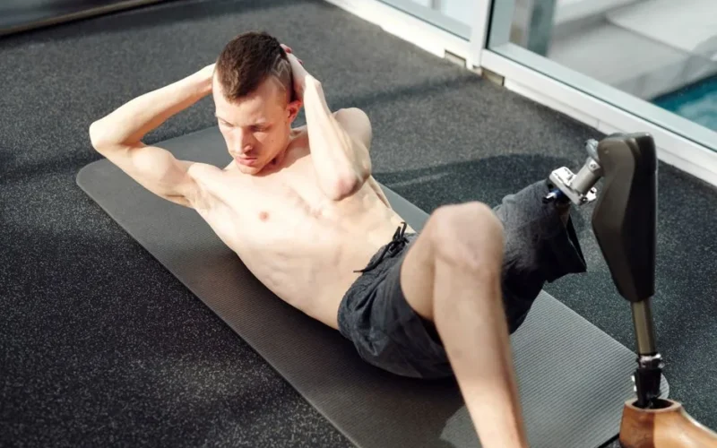 Determined man with prosthetic leg doing sit-ups on a yoga mat in a gym.