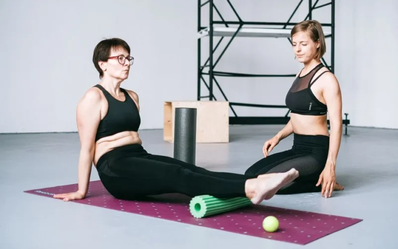 Two women in sportswear exercise with foam rollers on yoga mats in a gym environment.