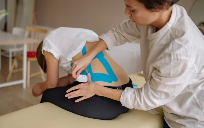 A therapist applies blue kinesiology tape to a patient's back in a clinical setting.