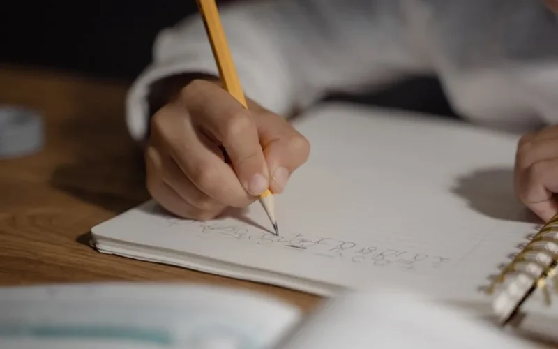 Close-up of a child writing homework using a pencil on a spiral notebook. Ideal for education themes.