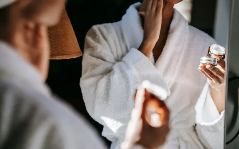Crop anonymous female in white bathrobe applying moisturizing facial cream on face while standing in front of mirror in bathroom
