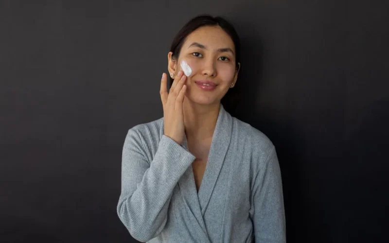 Positive ethnic female in bathrobe applying moisturizing cream on cheek during daily beauty routine against black background and looking at camera