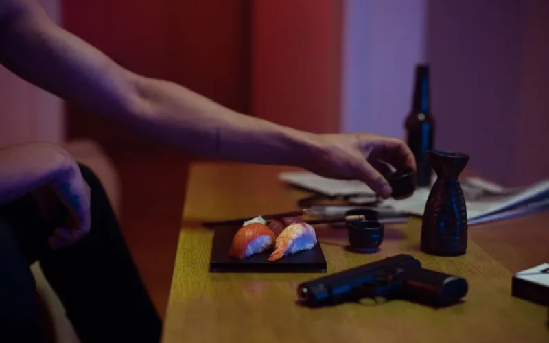 An intriguing scene with sushi, a gun, and sake glasses on a table under warm light.
