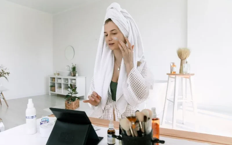 A woman in a bathrobe applies skincare products in a bright bathroom setting.