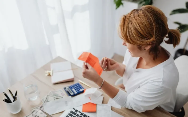 A woman organizing her finances with bills and receipts at a home office desk using a calculator and laptop.
