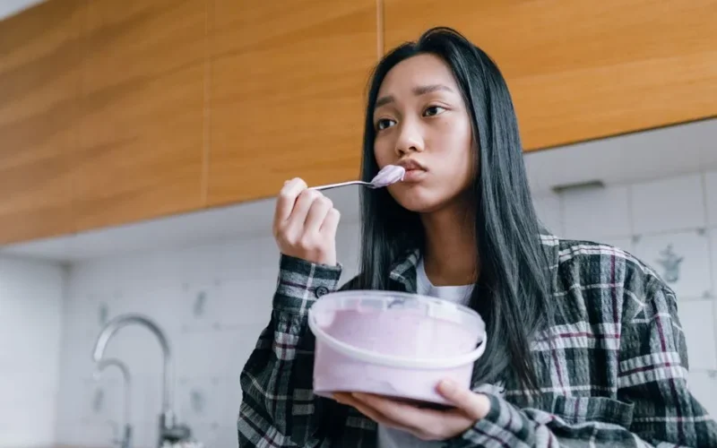 Asian woman enjoying ice cream in a casual indoor setting, looking pensive.