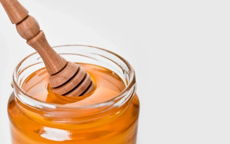 Glass jar of honey with a wooden dipper, set against a white background. Perfect for food and kitchen-related content.