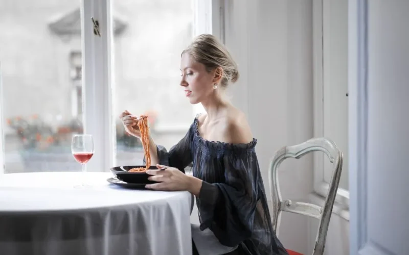 A stylish woman savoring spaghetti with red wine by a bright window.