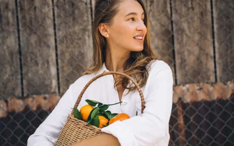 A cheerful woman in a white blouse holds a basket of ripe tangerines outdoors.