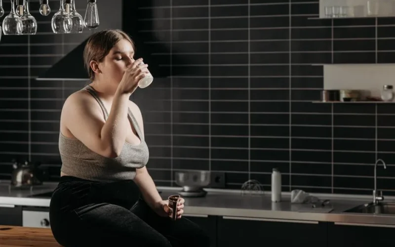Woman sitting on kitchen counter drinking milk in a modern setting.