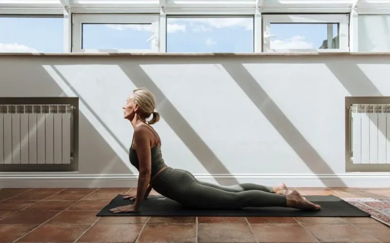 Senior woman performing yoga pose in a sunlit room, promoting healthy lifestyle and mindfulness.