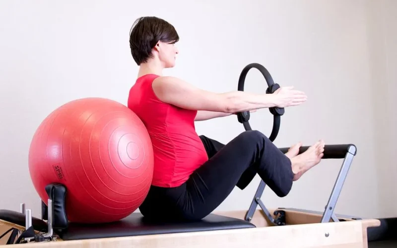 Pregnant woman in gym doing Pilates exercise with reformer and stability ball.