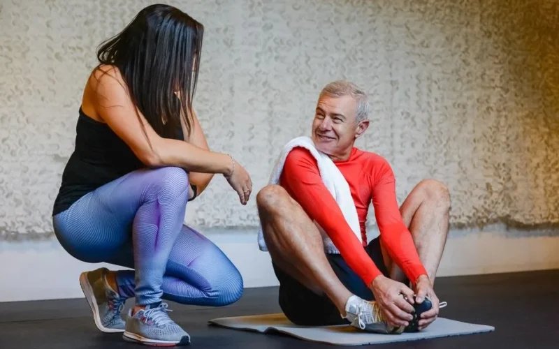 Fitness coach helps senior man stretch on exercise mat in a gym setting, promoting health and wellness.