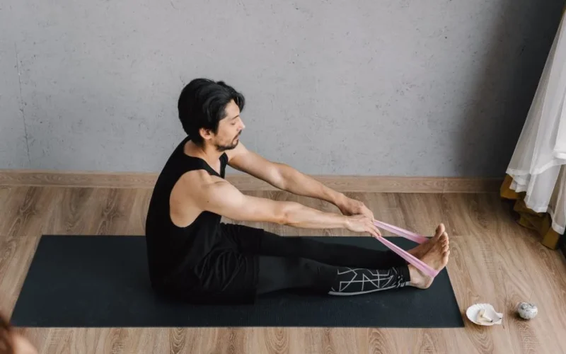 Man in black tank top performs yoga stretches with resistance band indoors.