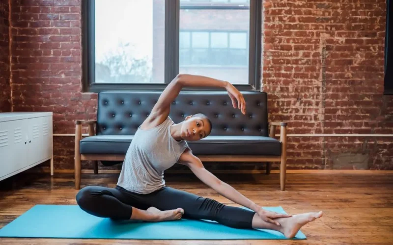 Full body of slim African American graceful female doing lateral bend exercise sitting on sports mat during training
