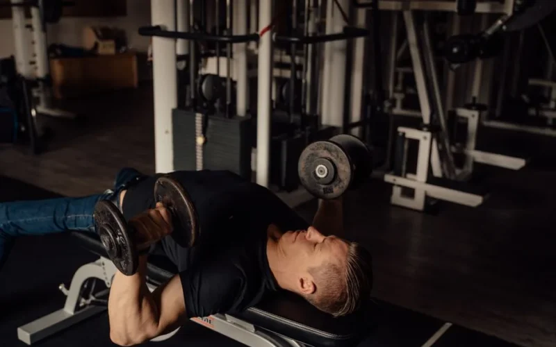 Muscular man doing dumbbell chest press exercise in a gym setting, highlighting fitness and strength.