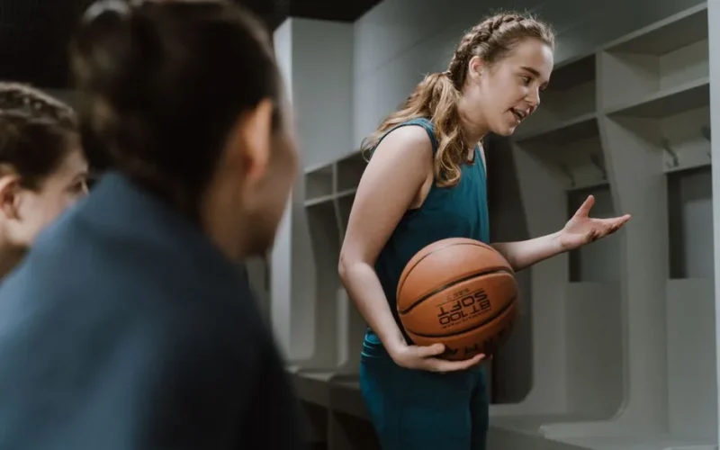 A young female basketball player explains strategies to teammates in a locker room.