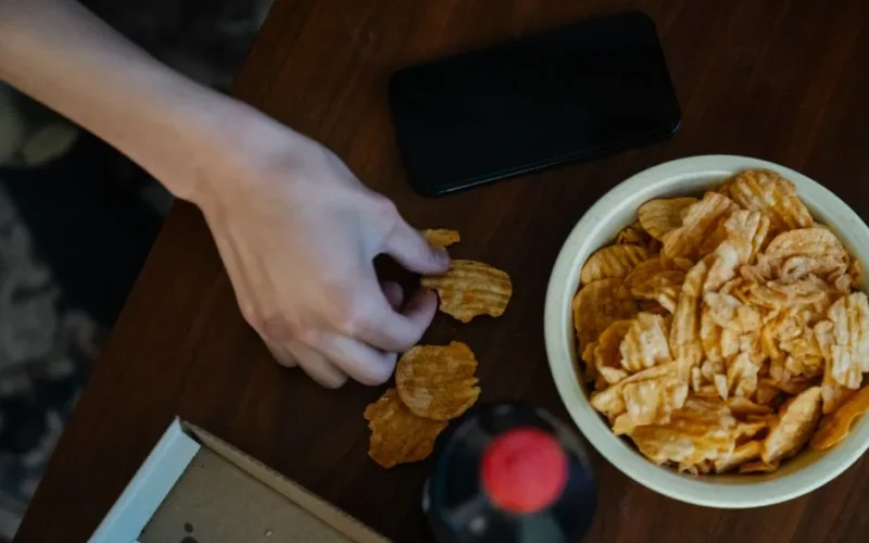 From above of crop anonymous person with crispy potato chips at table against bottle of refreshing drink and smartphone