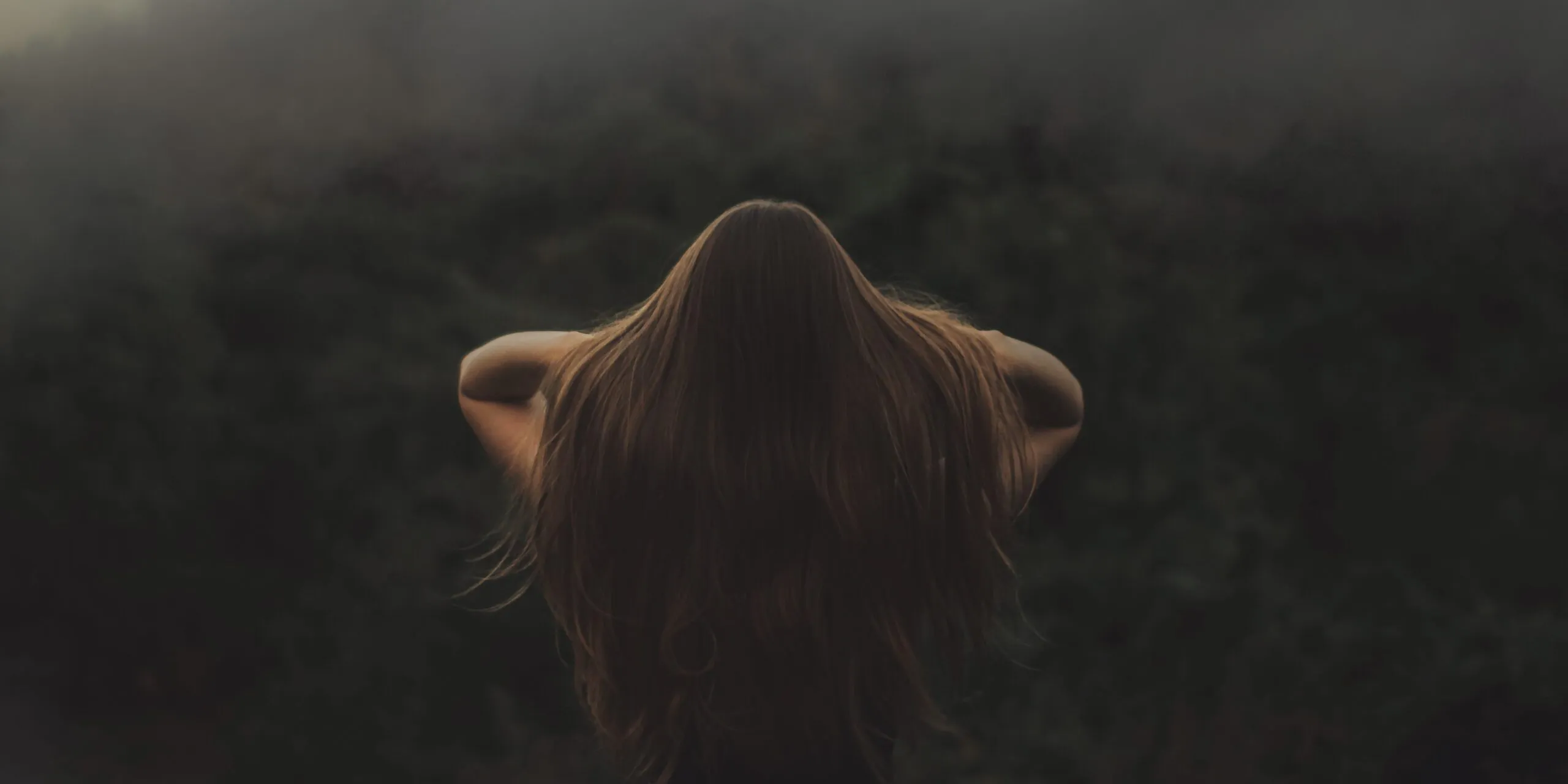Back view of a woman with long hair, standing in a misty forest.