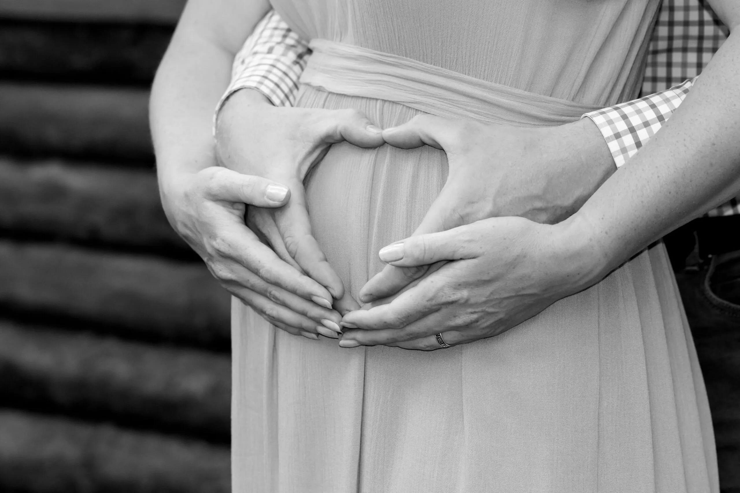 A loving couple forms a heart shape over a pregnant belly in a black and white portrait.