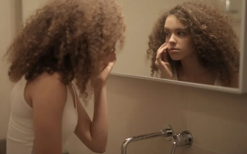 Teenage girl with curly hair examines her face in the bathroom mirror, reflecting contemplation.